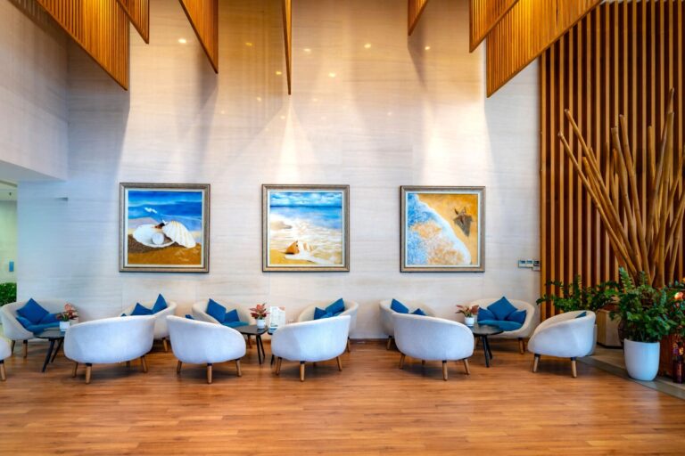 Bright double-height lobby lounge in a shared building common area, with rows of clean white armchairs, framed coastal artwork on a pale stone wall and a polished wood floor.
