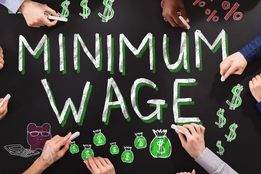 Chalkboard illustration of the words MINIMUM WAGE surrounded by chalk-drawn dollar signs, money bags and percent signs, with several hands drawing around them.
