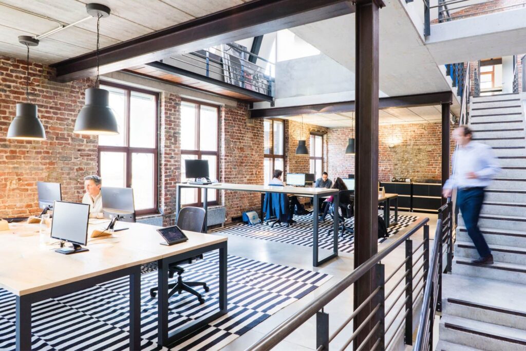Open-plan loft office with exposed brick walls, tidy shared desks and clean floors, and a staff member walking up the stairs.